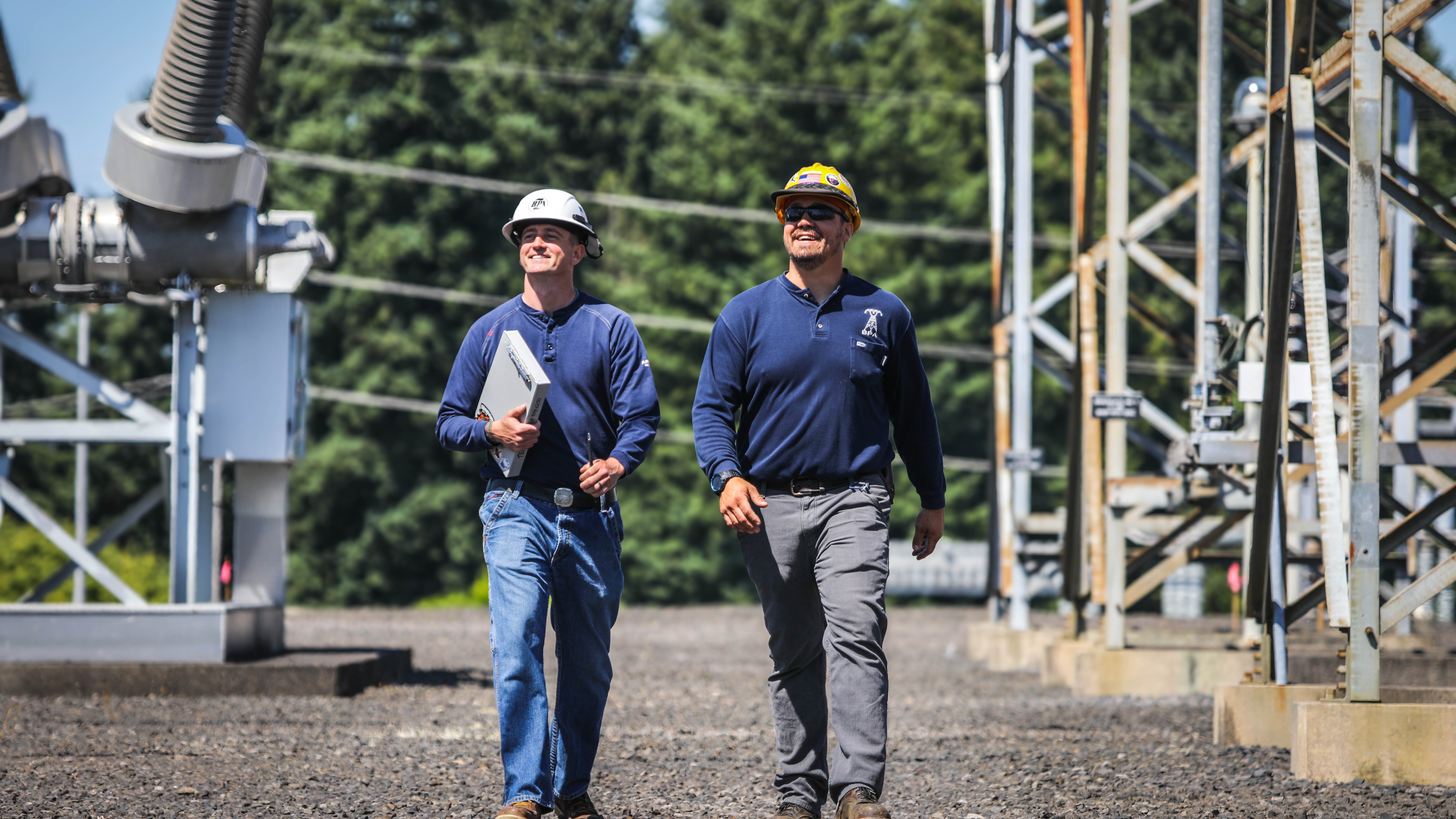 two men wearing hardhats, walking on gravel path under electrical equipment