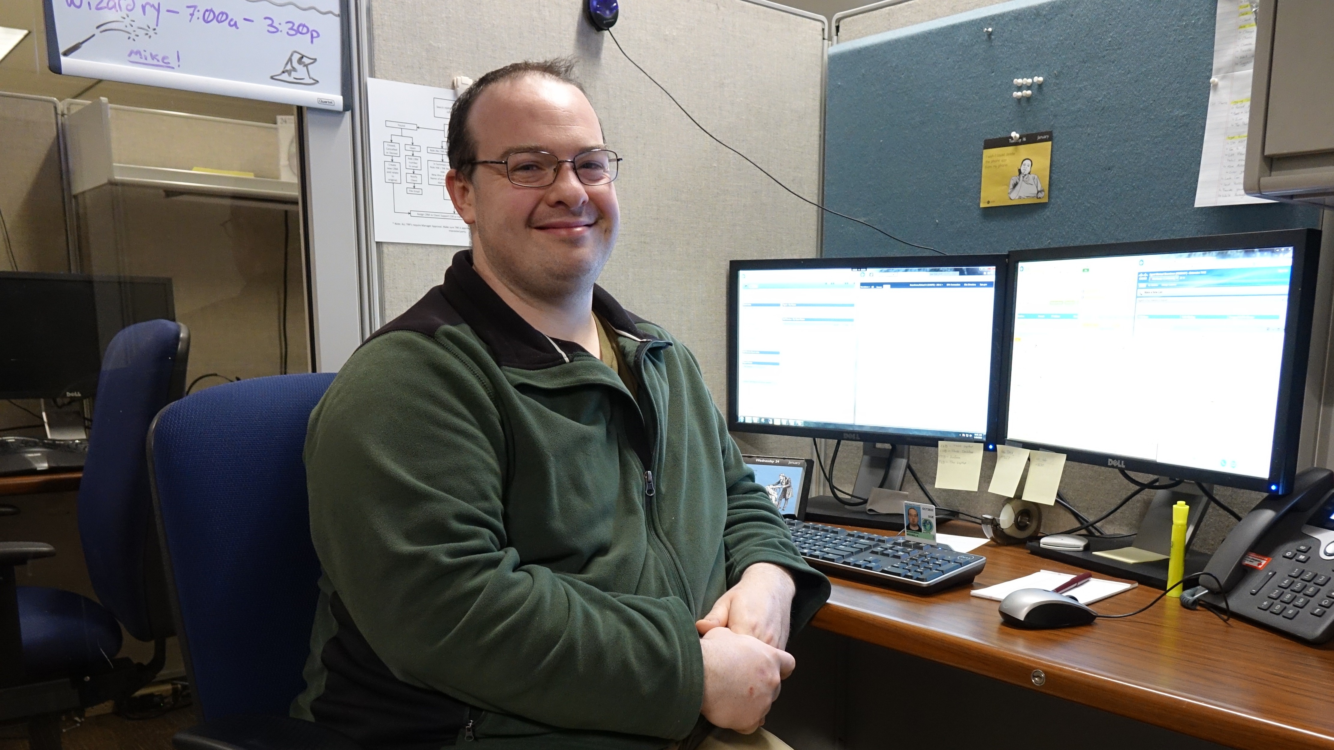 man sitting in front of a desk with two monitors on it, turned toward the camera and smiling
