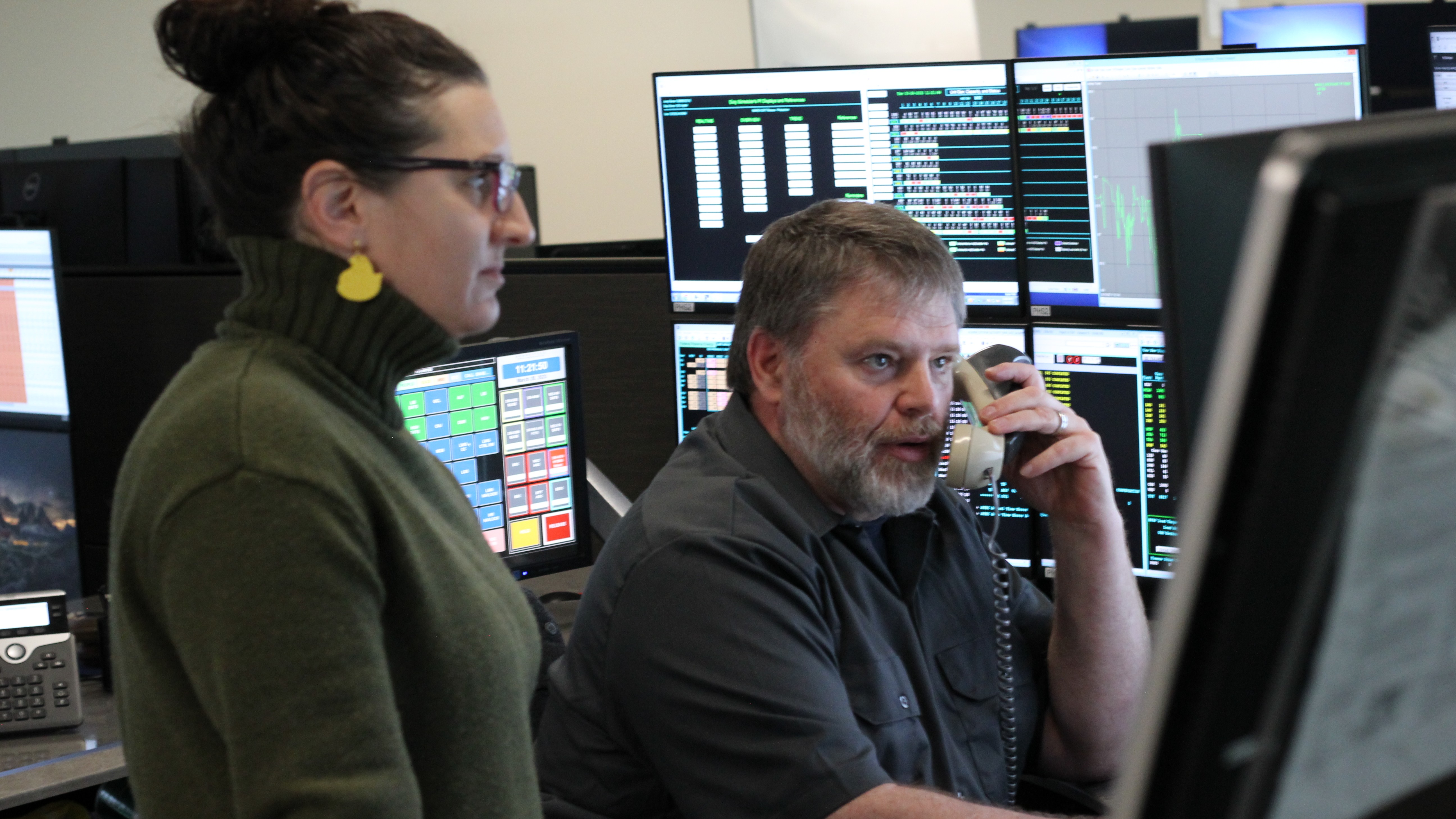 woman standing next to a man sitting at a desk, both looking at a monitor, while he holds a phone receiver