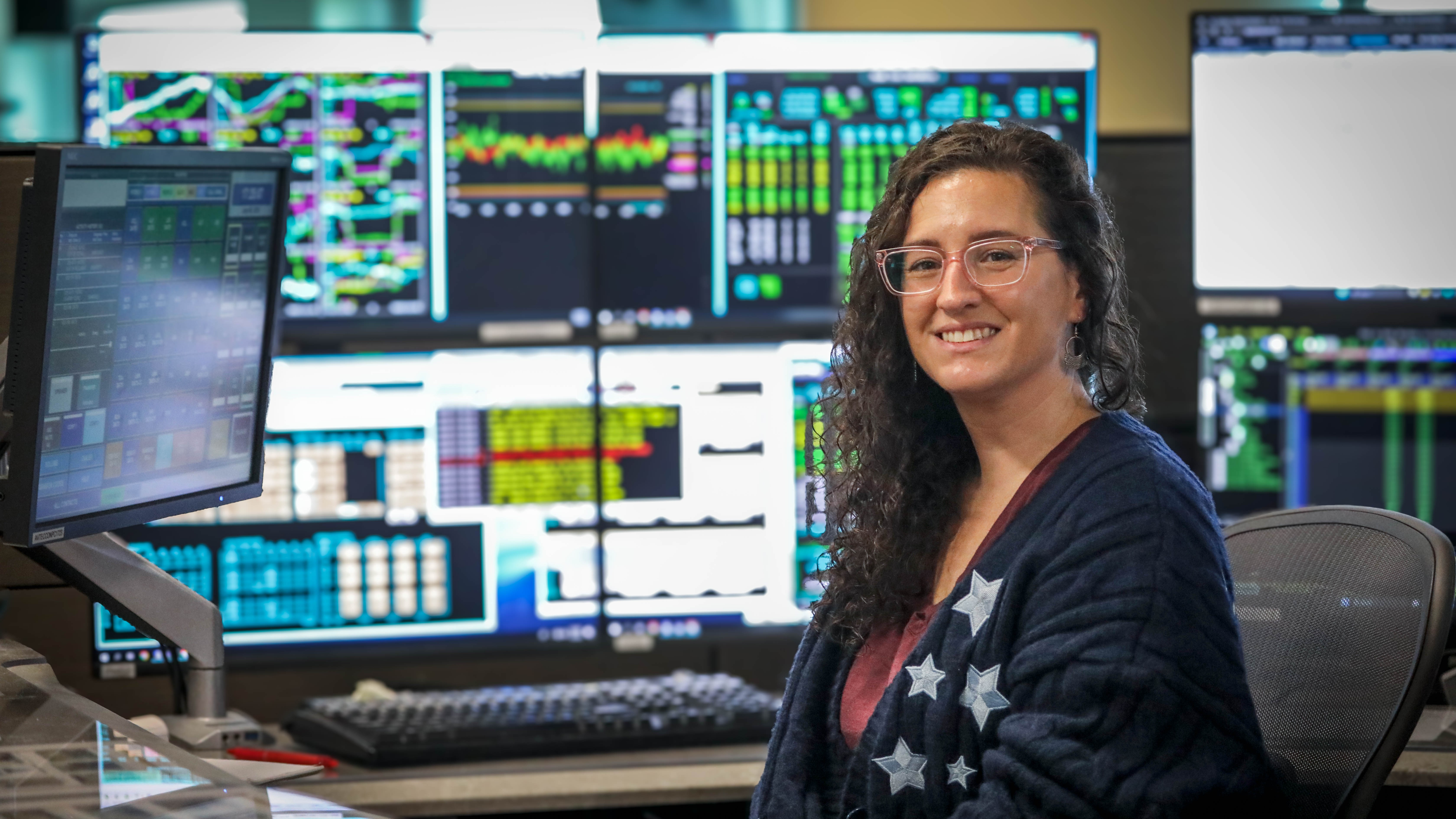 woman sitting at a desk, with monitors behind her, turned toward the camera and smiling