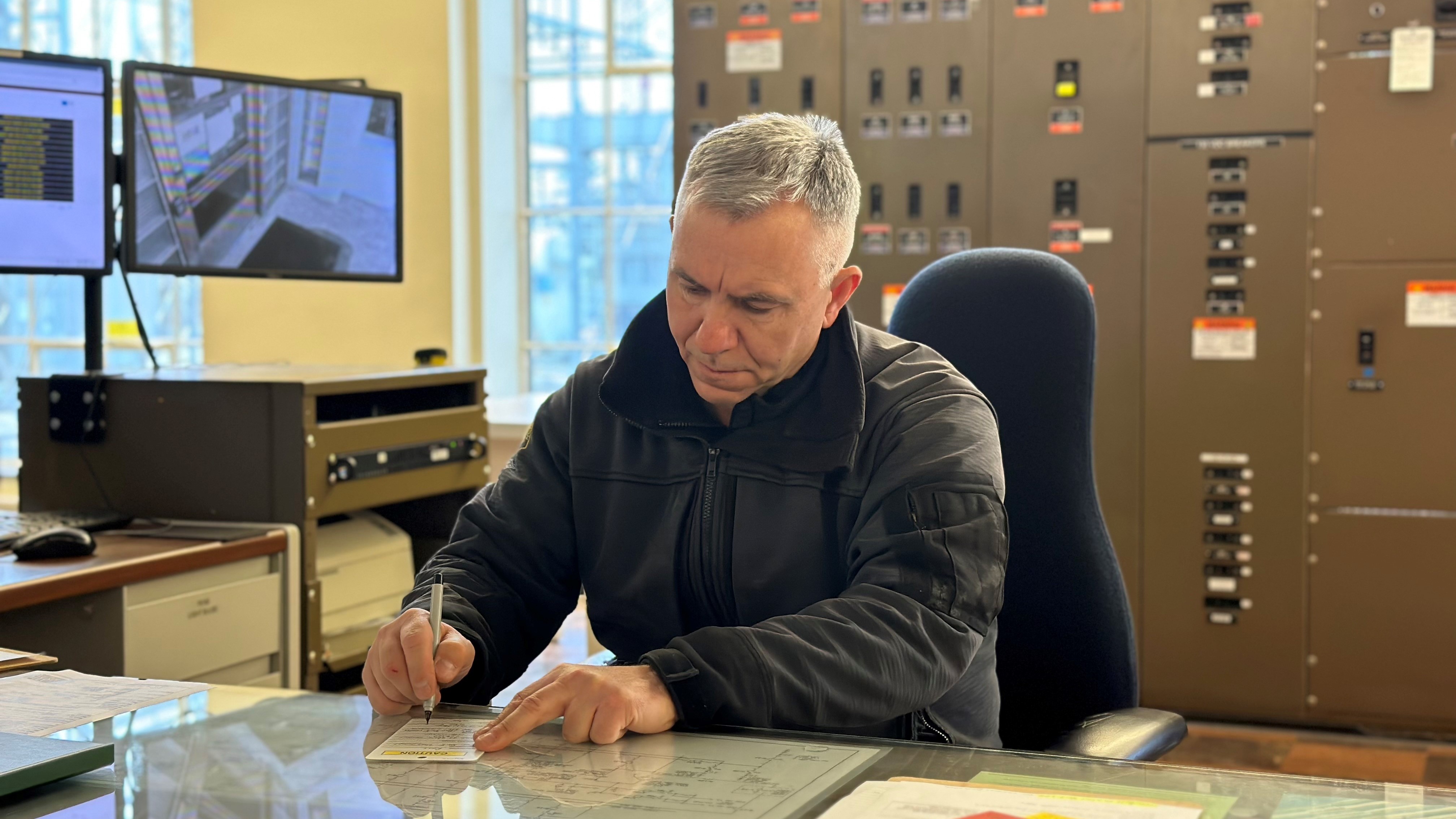 man sitting at a desk with monitors and electrical equipment behind him, writing on a document