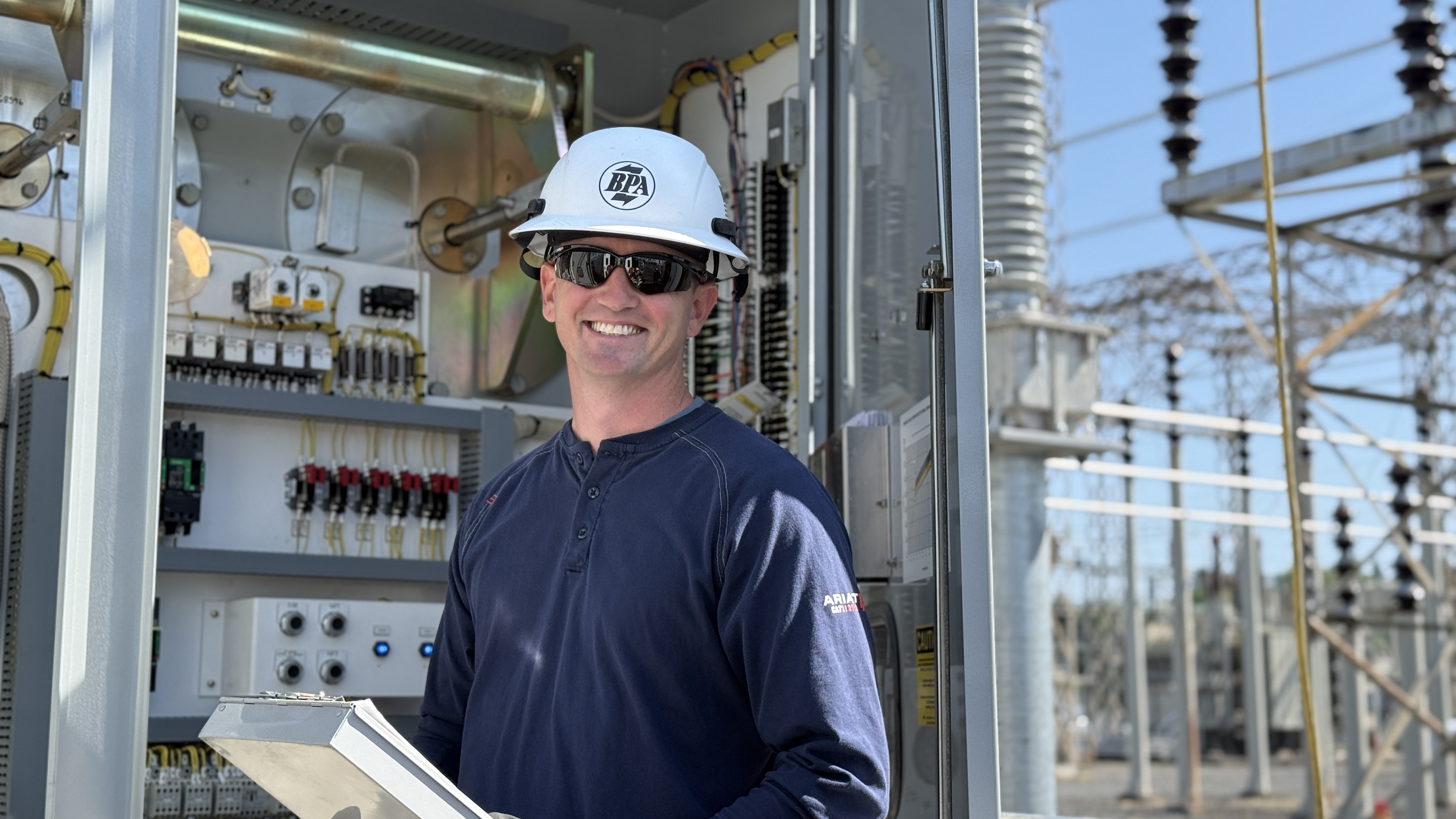 man wearing a hardhat and sunglasses, holding a clipboard, standing in front of electrical equipment outside and smiling at came