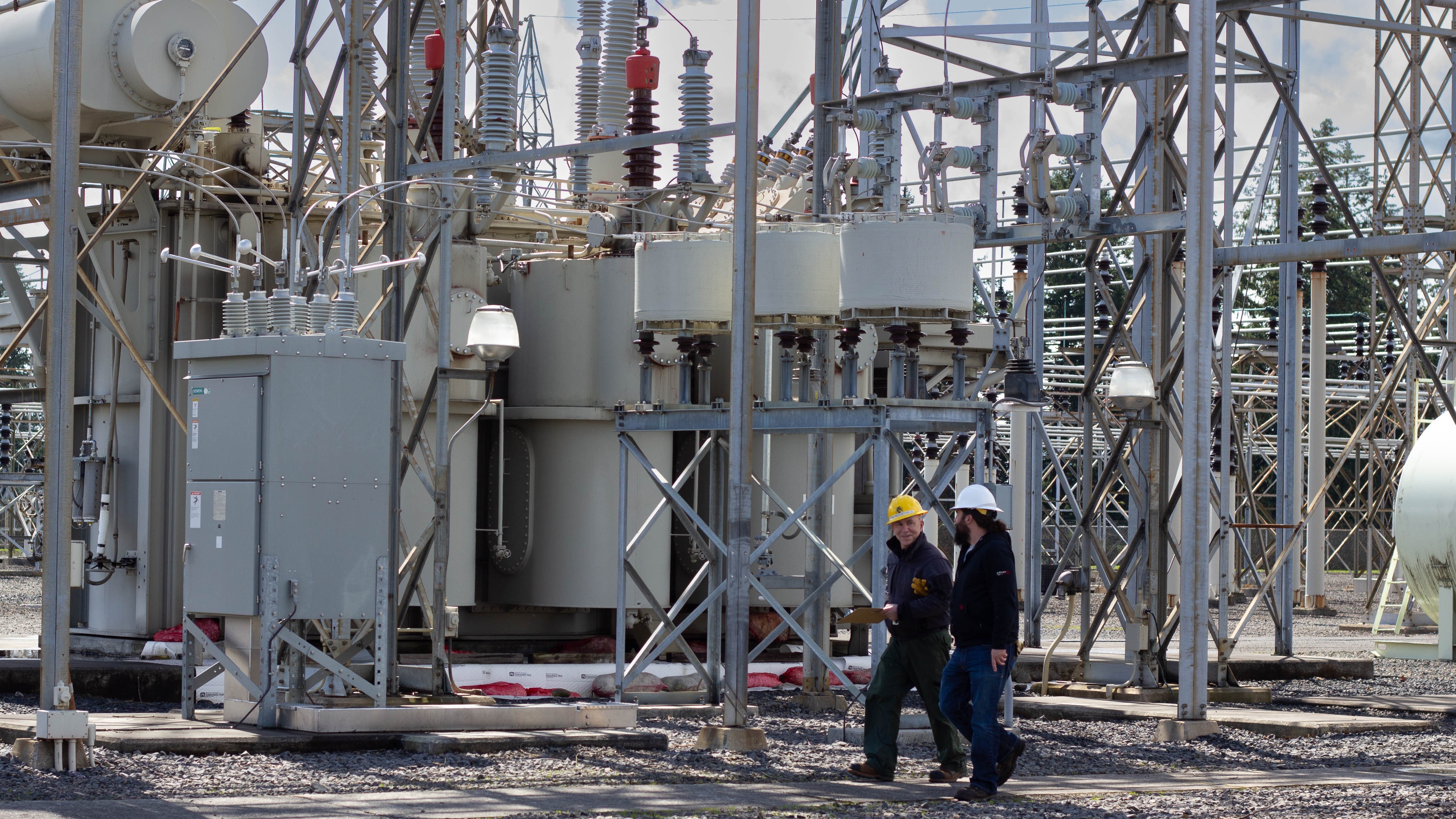two men wearing hardhats, walking in front of electrical equipment outside