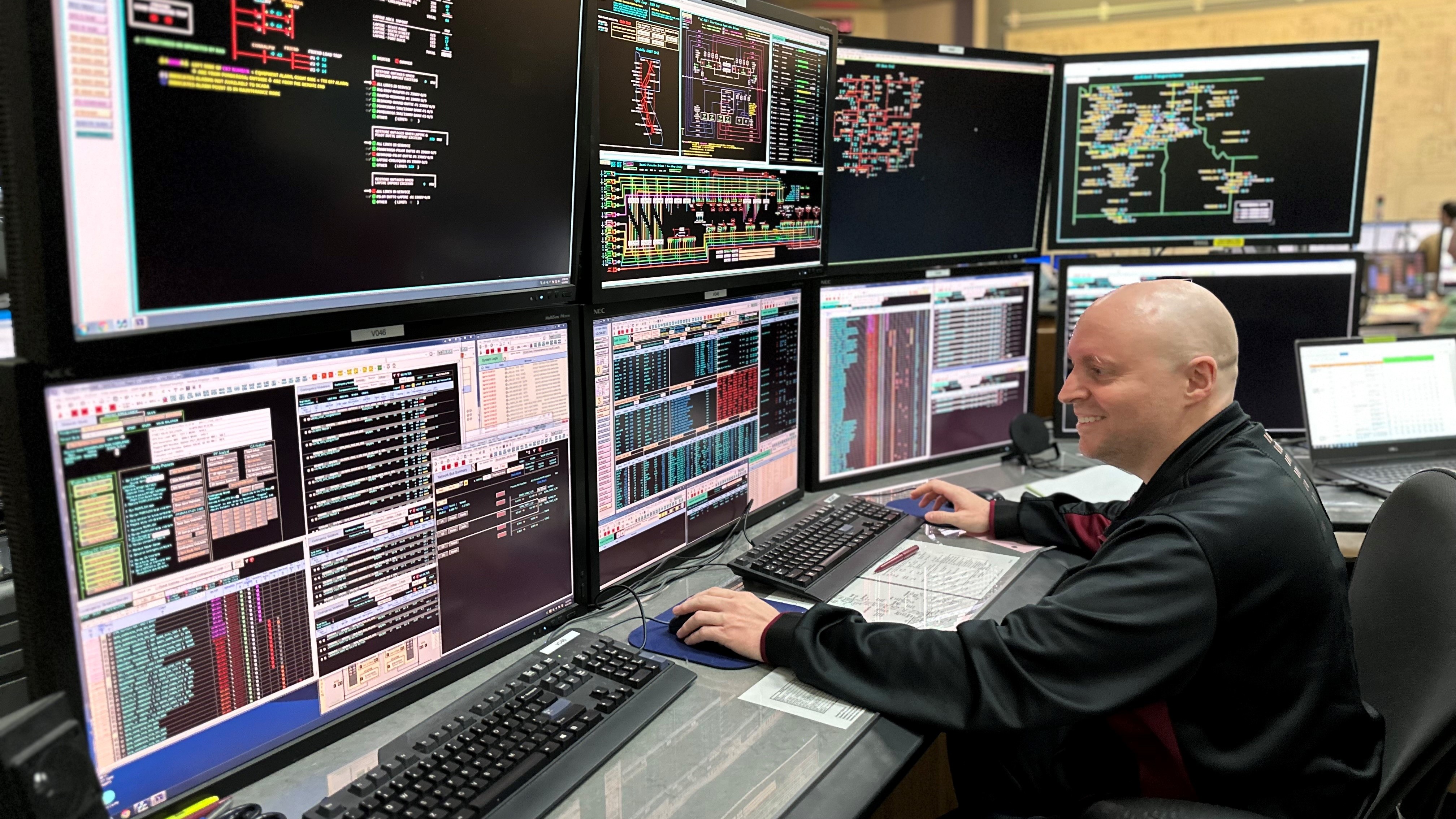 man sitting in front of eight monitors with colorful writing on the screens, with his hand poised above a mouse