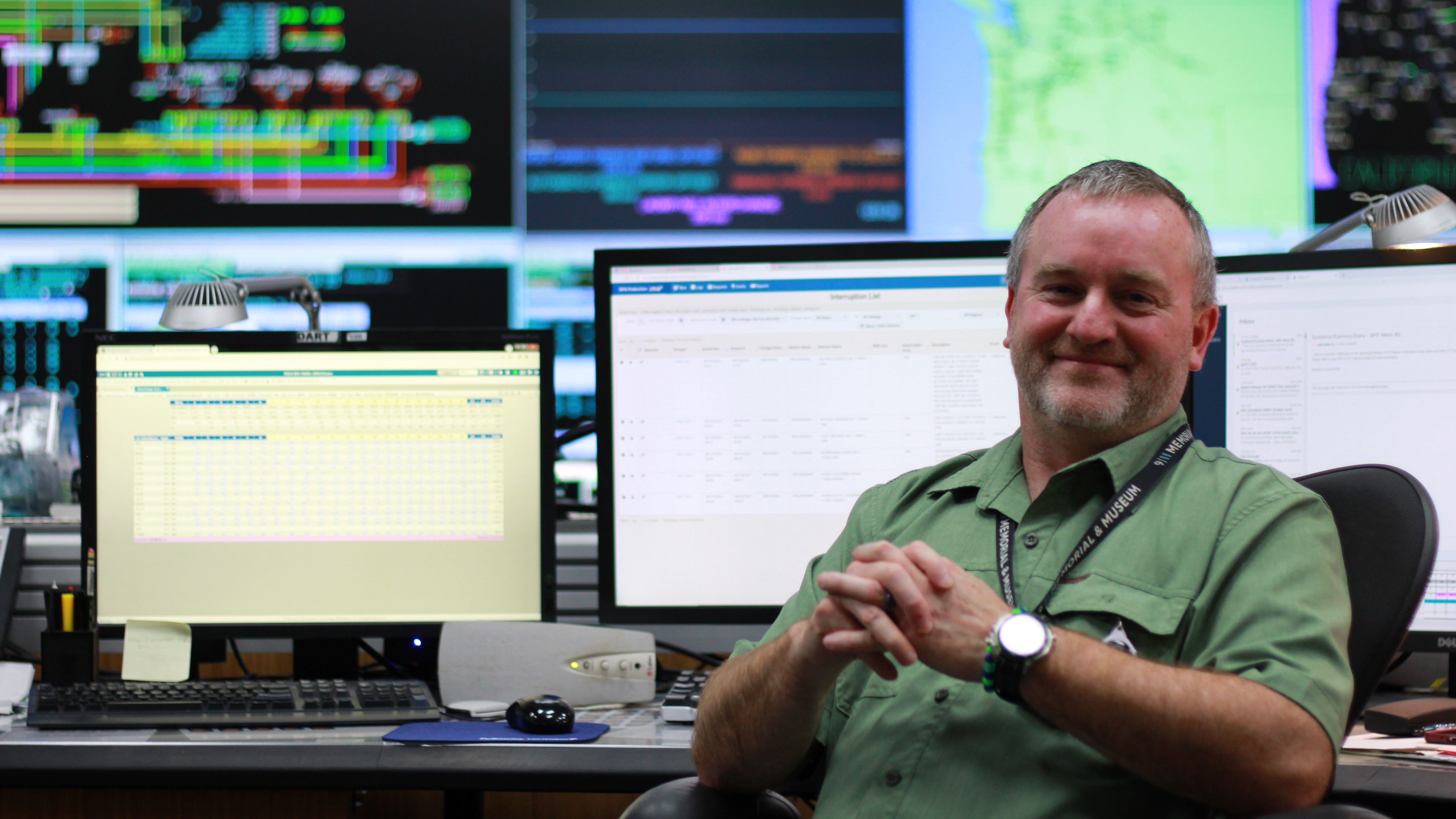 man sitting in a chair, swiveled away from a desk with multiple monitors, smiling at the camera