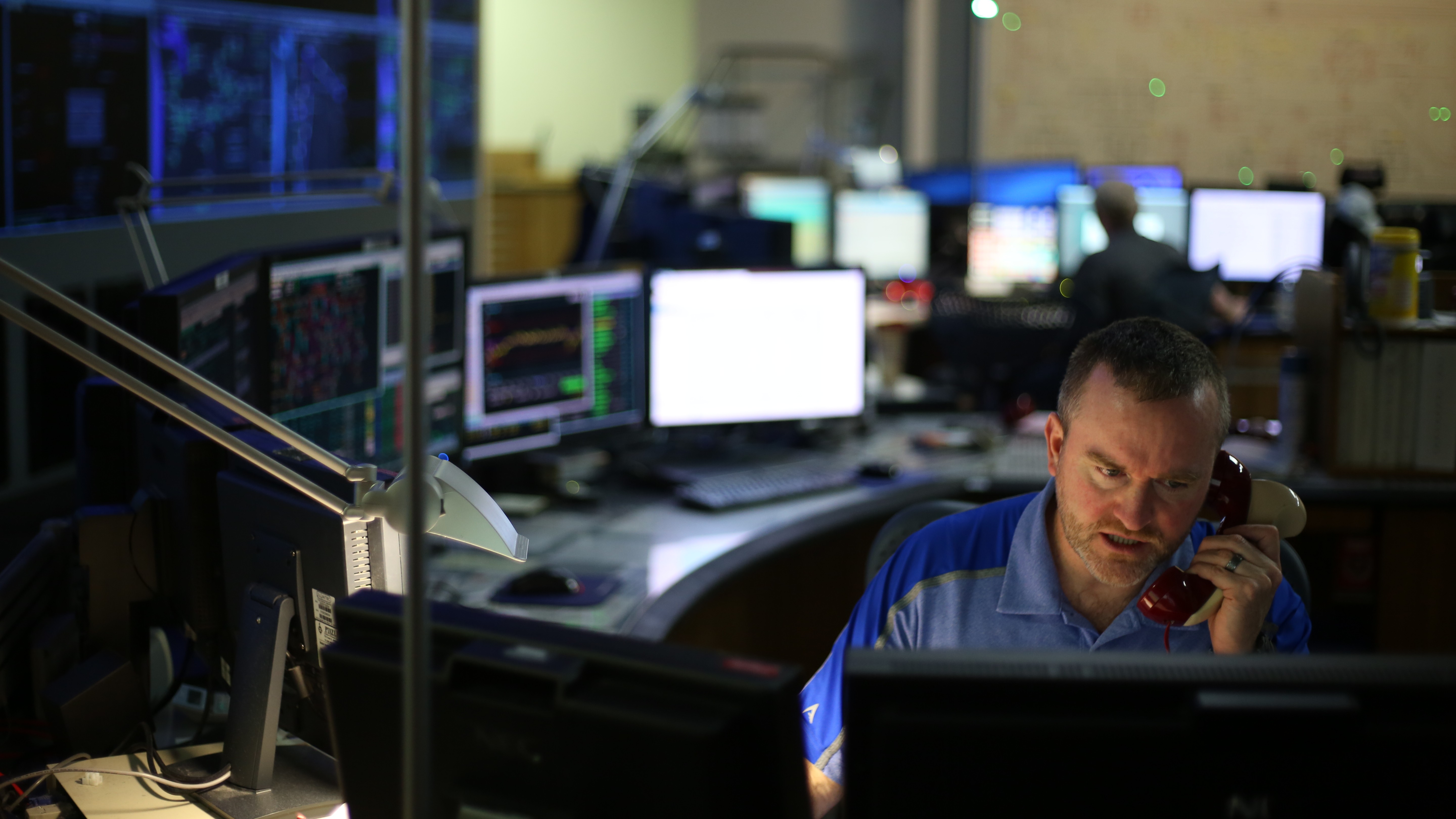 man sitting a desk in dark, in front of many monitors, holding a phone