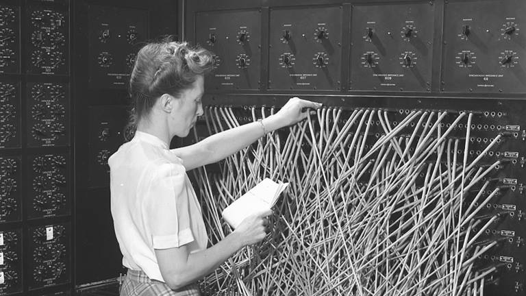 woman standing a room consumed by large computing device, holding a piece of paper while touching the computer