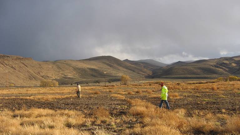 two people walking through a desolate, high-desert landscape filled with yellow grass and brown hills in the background