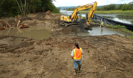 person with orange vest standing in mud watching heavy equipment move dirt in giant puddle of water
