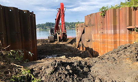 large piece of equipment digging in dirt with fences in foreground and water in background