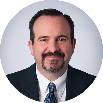 picture of man with dark brown hair, parted on the side, wearing a black suit with a white shirt and blue patterned tie, set against a grey background, photo in shape of circle