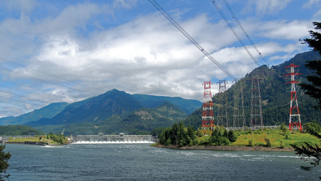 river with a concrete structure spanning across it in the distance with transmission towers set on a green grassy shore with transmission lines spanning across the water, blue mountains in the background 