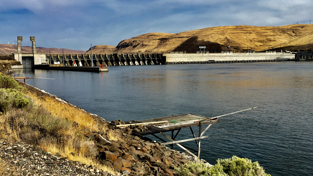 water with a rocky shore in the foreground, olive colored hilly terrain on the other side of the water and spanning across the water a concrete structure